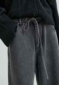 Tuvumā tumši pelēkie džinsi ar sasietu auklu viduklī, valkāti ar melnu ribotu adījuma džemperi ar lielu pogu.