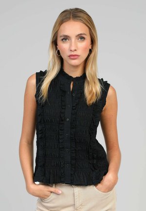 Femme blonde avec des boucles d'oreilles créoles portant un chemisier noir sans manches à volants et boutons, pantalon beige, mains dans les poches, fond neutre.