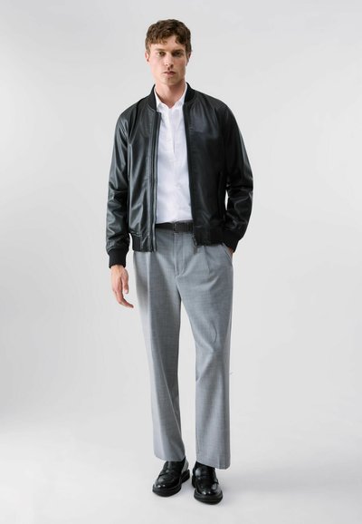 Jeune homme portant une veste en cuir noire, une chemise blanche, un pantalon gris et des chaussures noires, debout devant un fond clair uni.