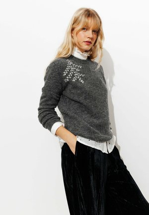 Pull en laine gris avec des ornements sur l'épaule gauche, superposé à une chemise blanche à col, associé à un pantalon en velours noir.