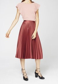 Donna che indossa una blusa rosa a maniche corte e una gonna midi plissettata a vita alta in mauve con scarpe nere con lacci.