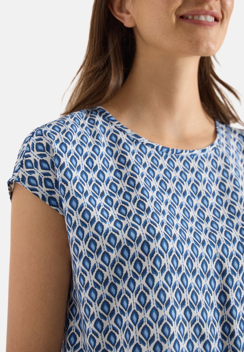 Femme portant un chemisier à manches courtes avec un motif géométrique bleu et blanc, souriante, avec de longs cheveux bruns visibles sur fond blanc.