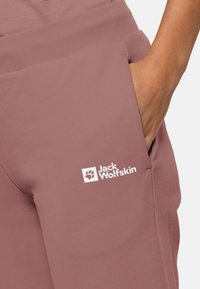 Person’s hand in pocket of mauve pants with Jack Wolfskin logo on upper thigh against a plain light background.