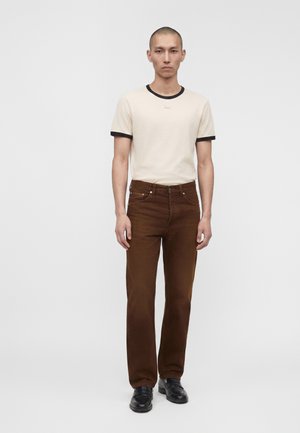 Homme debout devant un fond uni, portant un t-shirt crème avec bordure noire, pantalon marron et chaussures noires, regardant droit devant avec une expression neutre.