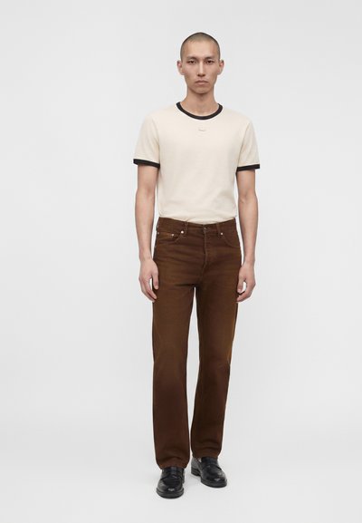 Homme debout devant un fond uni, portant un t-shirt crème avec bordure noire, pantalon marron et chaussures noires, regardant droit devant avec une expression neutre.