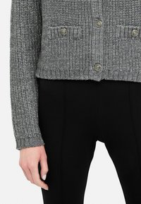 Cardigan grigio a maglia con una texture a costine, dotato di due tasche sul petto e dettagli in metallo sui bottoni. Indossato sopra pantaloni neri aderenti.