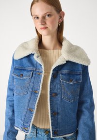 Dżinsowa kurtka w średnim odcieniu niebieskiego, z puszystym kremowym kołnierzem, dwiema kieszeniami na piersi i brązowymi guzikami. Postrzępiony dół oraz widoczne detale w szyciu.