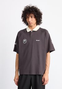 Polo de color gris oscuro con un color de collar crema, con un logo en el pecho, mangas cortas y un ajuste relajado.