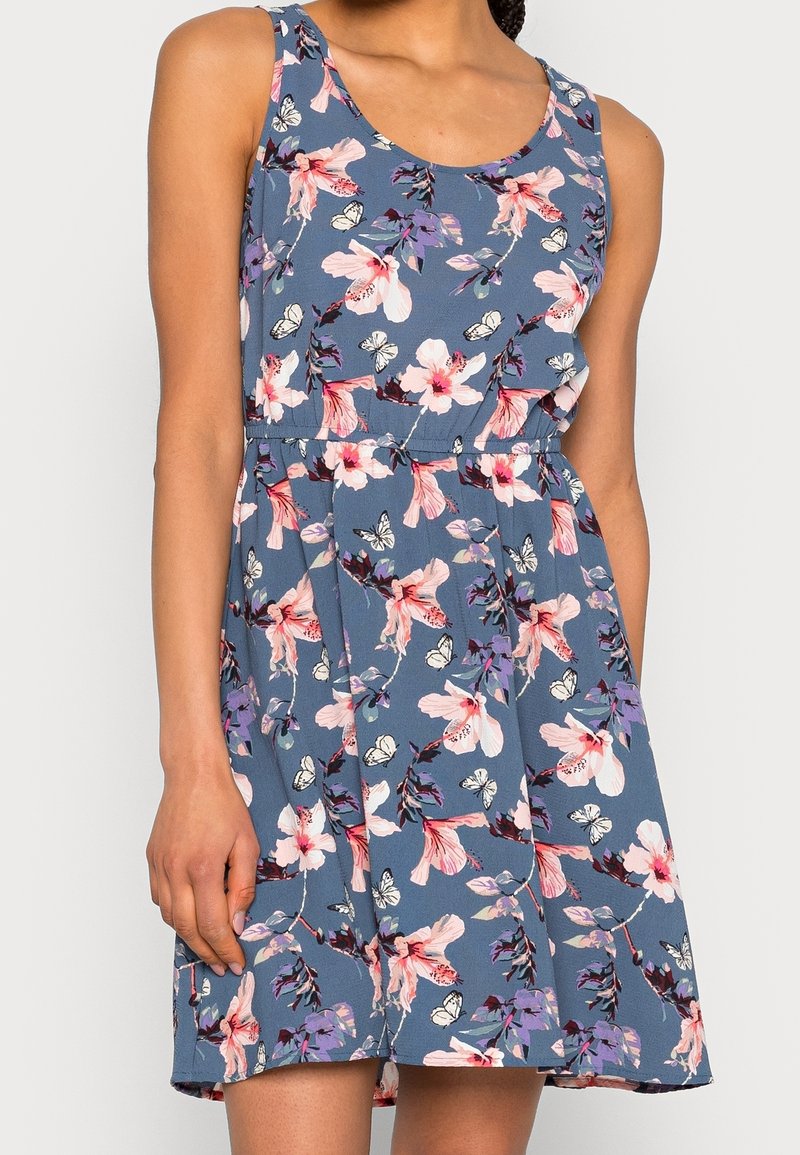 Robe bleue sans manches avec des motifs floraux roses et violets ainsi que des papillons blancs, portée par une personne debout devant un fond uni.