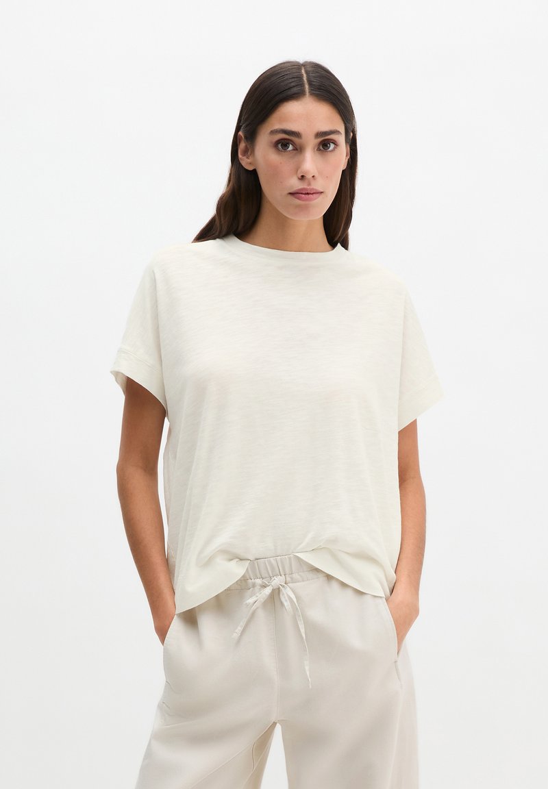 Femme aux longs cheveux foncés portant un t-shirt crème à manches courtes rentré dans un pantalon beige clair à cordon, debout les mains dans les poches.