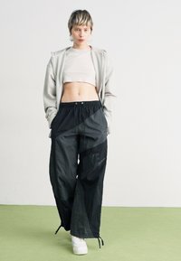 Sudadera con cremallera de color gris claro sobre una camiseta corta de color crema, combinada con pantalones de chándal negros holgados que presentan un gran logo y cordón. Zapatillas blancas.