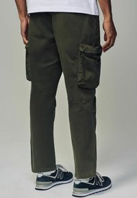 Pantaloni cargo de culoare verde măslin închis, cu multiple buzunare laterale, croială lejeră și asortați cu adidași negri și gri. Textură netedă.