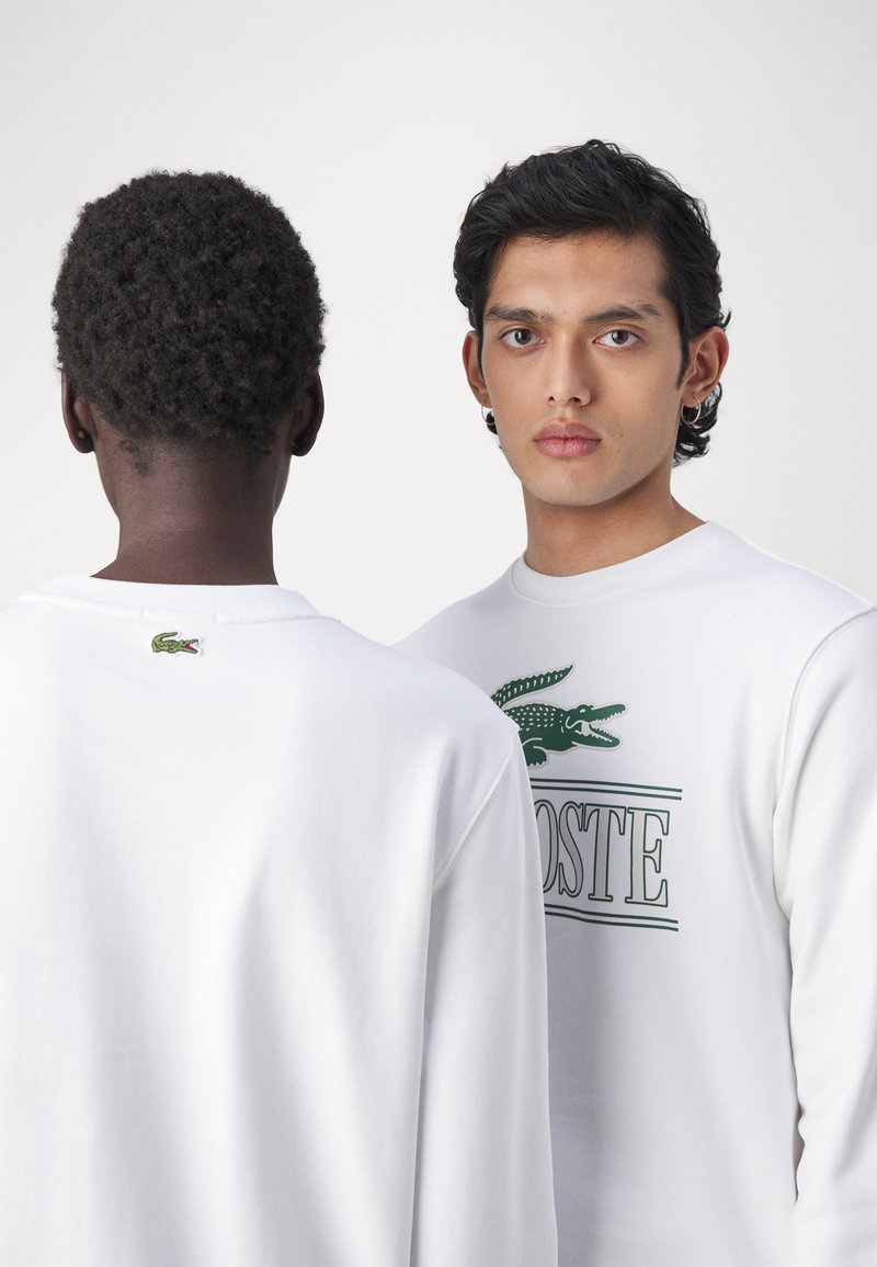 Sudadera blanca con logotipo verde de Lacoste y texto, hecha de una tela suave. Exhibida desde dos ángulos, enfatizando el diseño trasero y delantero.