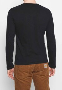 Schwarzes Langarmshirt mit Rundhalsausschnitt, aus glattem Stoff gefertigt. Kombiniert mit braunen Cordhosen mit Gesäßtaschen und einem Logopatch.