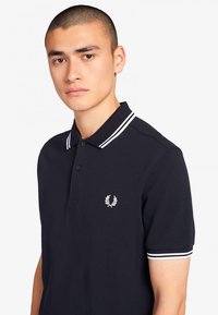 Polo de color navy hecho de algodón texturizado, con una tapeta de dos botones, acentos de rayas blancas en el cuello y las mangas, y un logo en el pecho.