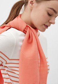 Koraalroze geribde sjaal rondom de nek, gemaakt van zachte stof, gecombineerd met een casual witte top met horizontale oranje gestreepte lijnen.