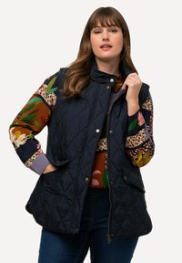 Navy farbener gesteppter Weste mit Stehkragen, ausgestattet mit Druckknopf-Details, seitlichen Taschen und einer glatten Textur über einem floral gemusterten Hemd.