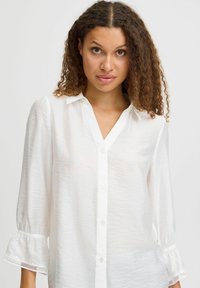White button-up shirt with a v-neck, long sleeves, gathered cuffs, textured fabric, and lace accents on the sleeves.