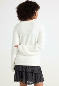 Un pull blanc duveteux à manches longues, associé à une jupe noire en dentelle à plusieurs couches avec des bords froncés. Texture lisse, coupe décontractée.