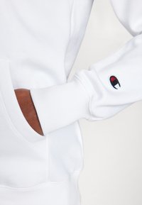 Hvit hettegenser med ribbestrikkede mansjetter og en frontlomme. Har en liten brodert logo med røde og svarte detaljer på ermet.
