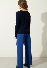 Eine Frau, die mit dem Rücken zur Kamera steht, trägt ein navyblaues langärmliges Oberteil, leuchtend blaue weit geschnittene Hosen und schwarze Schuhe vor einem schlichten hellen Hintergrund.