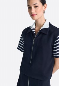 Tengerészkék cipzáras felső fehér gallérral és csíkos rövid ujjakkal. A szövet texturált hatású, testhezálló szabású, kontrasztos varrással.