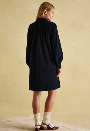 Marineblaues Cordkleid mit langen Ärmeln, gerafftem Schulterbereich und entspannter Silhouette, kombiniert mit cremefarbenen Socken und burgunderroten Schuhen.