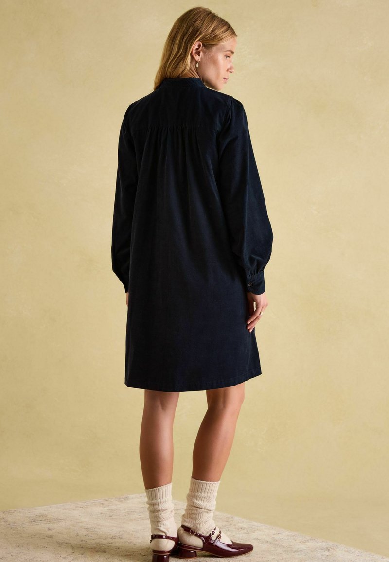 Marineblaues Cordkleid mit langen Ärmeln, gerafftem Schulterbereich und entspannter Silhouette, kombiniert mit cremefarbenen Socken und burgunderroten Schuhen.