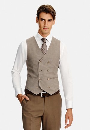Mężczyzna noszący beżową dwurzędową kamizelkę, białą koszulę, brązowy krawat w groszki i brązowe spodnie, z jedną ręką w kieszeni.