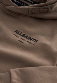 Sweat à capuche marron avec logo noir "ALLSAINTS", caractères japonais et "LONDON" imprimés sur la poitrine, montrant le cordon de serrage et la texture du tissu.