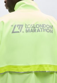 Άτομο που φοράει ανοιχτό πράσινο μπουφάν με ανακλαστικές ρίγες και την επιγραφή "tcs London Marathon 2026" σε γκρι στο πίσω μέρος.