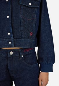 Giacca di jeans blu scuro con bottoni a pressione, tasche sul petto e cuciture rosse. Abbinata a jeans blu scuro coordinati con una cintura con logo.