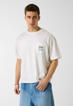 Młody mężczyzna w białej koszulce z krótkim rękawem z logo na kieszeni na piersi z napisem "Malibu Guess Jeans 1981" oraz jasnoniebieskich dżinsach.