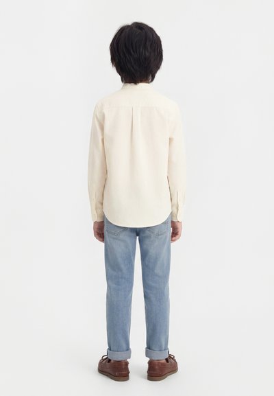 Παιδί που κοιτάζει προς τα πίσω, φοράει μια κρέμ μπλούζα με μακριά μανίκια, ανοιχτό μπλε τζιν με γυρισμένα μπατζάκια και καφέ παπούτσια, στέκεται μπροστά σε απλό λευκό φόντο.