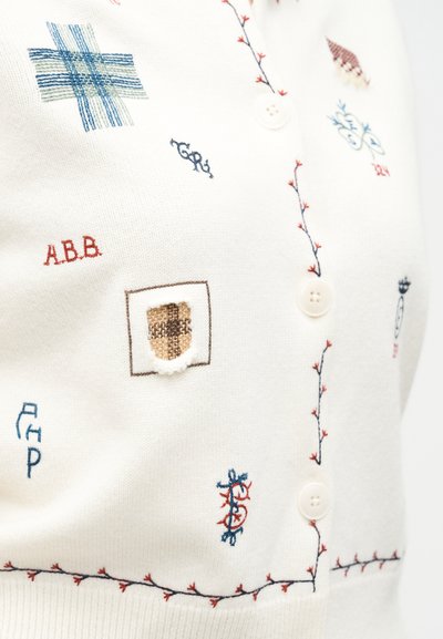 Λευκό ζακέτα με μικρά πολύχρωμα κεντήματα, κουμπιά και κόκκινη και μπλε ραφή σε σχήμα κλήματος κατά μήκος της γραμμής των κουμπιών και του τελειώματος.