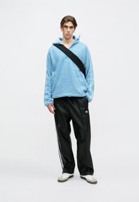 Giovane in felpa di pile azzurro chiaro, pantaloni della tuta neri Adidas, sneakers bianche e borsa a tracolla nera, in piedi di fronte a uno sfondo semplice.