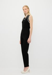 Débardeur noir avec des bordures blanches, associé à un pantalon noir ajusté. Modèle portant des talons blancs, debout devant un fond uni.