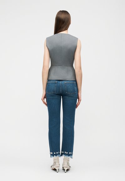 Γυναίκα με μακριά καστανά μαλλιά, φορεί γκρι αμάνικη μπλούζα με πεπλούμ, μπλε τζιν με floral λεπτομέρειες στο τελείωμα και λευκά σανδάλια με τακούνι, στέκεται γυρισμένη πλάτη.