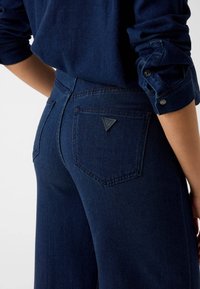 Un jean en denim foncé présente un dos ajusté avec un patch logo triangulaire sur la poche droite et une finition texturée subtile.