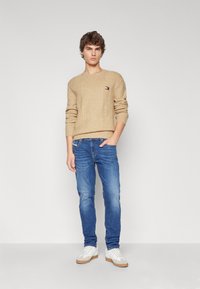 Beige stickad tröja med kabelmönster och liten logotyp, tillsammans med blå denimjeans och vita sneakers med beige detaljer.