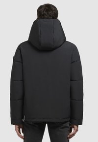 Muškarac u crnoj zimskoj jakni s velikim kapuljačom, prikazan sa stražnje strane na jednostavnoj sivoj pozadini.