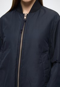 Tamsiai mėlynas bombonešio stiliaus švarkelis su aukštu apykakliu, auksine užtrauktuku ir šoninėmis kišenėmis; pasižymi lygia tekstūra ir subtiliomis siuvimo detalėmis.