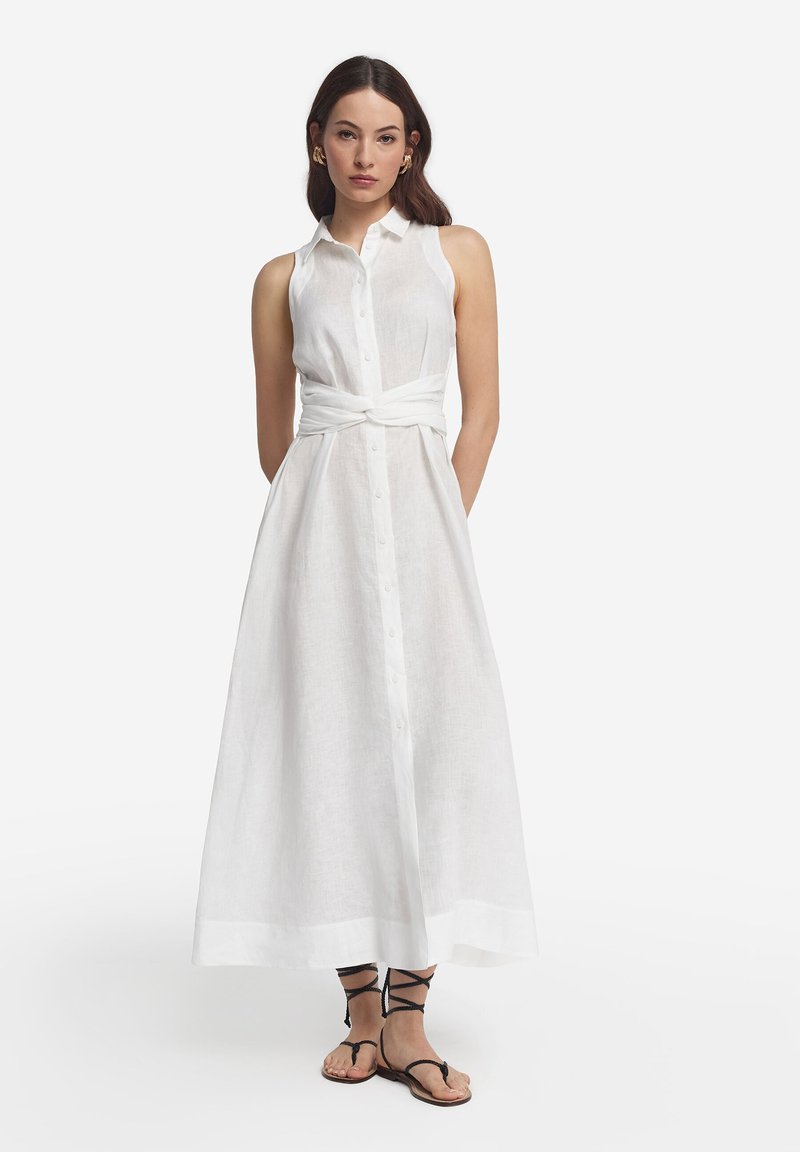 Femme portant une robe maxi blanche sans manches boutonnée avec une ceinture à la taille et des sandales noires à lacets, debout devant un fond uni.