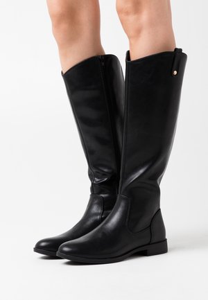 Botas de piel negra hasta la rodilla con una textura suave, cierre de cremallera lateral, puntera redondeada y suela plana de goma. Presenta un pequeño acento metálico.