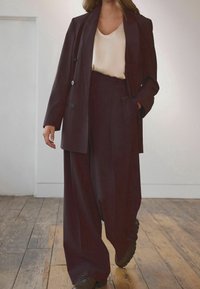 Costume pantalon bordeaux avec un blazer croisé et un pantalon à jambes larges. Haut beige en satin en dessous, associé à des chaussures marron. Fond en parquet en bois.
