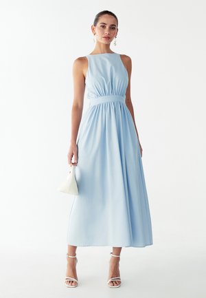 Lichtblauwe mouwloze jurk met een geplooide taille, een vloeiende rok en een gladde textuur. Het model houdt een kleine witte handtas vast en draagt sandalen met bandjes.