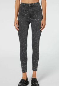 Jeans skinny de mezclilla negra con detalles de tachuelas plateadas, diseño de cintura alta y una textura suave, acabados con un cierre de cremallera y botón.