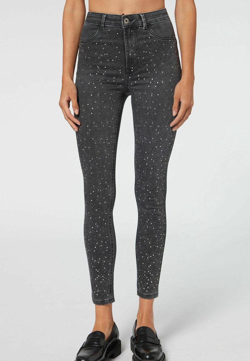 Jeans skinny de mezclilla negra con detalles de tachuelas plateadas, diseño de cintura alta y una textura suave, acabados con un cierre de cremallera y botón.