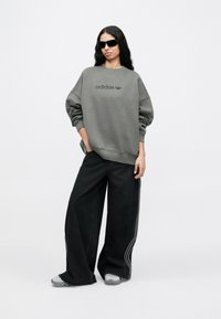 Γκρι υπερμεγέθη φούτερ με λογότυπο adidas σε τονικά χρώματα, συνδυασμένο με μαύρο τζιν παντελόνι φαρδιάς γραμμής με ραφές στο πλάι. Το μοντέλο φοράει μαύρα γυαλιά ηλίου.