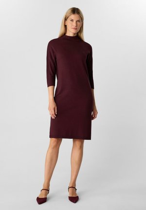 Woman standing in a knee-length, dark burgundy dress with three-quarter sleeves and matching pointed-toe heels against a plain background.